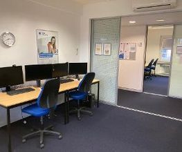 Offener Lernbereich mit vier PC-Arbeitsplätzen mit jeweils 2 Bildschirmen, einem Bürostuhl, Tastatur, Maus und Headset. An der Wand eine Uhr und ein Plakat.