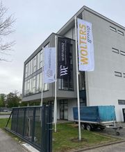 mediaService Bremen | Full-Stack Werbeagentur - Drei große weiße und schwarze Werbefahnen an Fahnenmasten wehen vor einem modernen Bürogebäude mit angeschlossenem Metalltor.