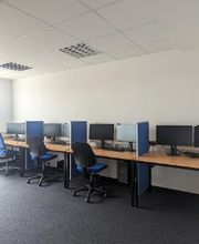 Schulungsraum mit mehreren Arbeitsbereichen, die durch blaue Trennwände voneinander abgegrenzt sind und mit PC-Bildschirmen, Tastatur, Maus und Headset ausgestattet sind. Blaue Bürostühle.