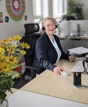 Frau sitzt am Arbeitsplatz mit Tastatur und Maus. Neben ihr ein gelber Blumenstrauß, hinter ihr ein großes Plakat