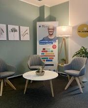 Eingangsbereich mit grüner Wand an der 3 Bilder mit Pflanzen hängen. Im Raum steht ein Beistelltisch mit 3 grauen Sesseln. Dahinter steht ein Banner und eine Stehlampe. Rechts ein Kleiderständer und eine Pflanze.