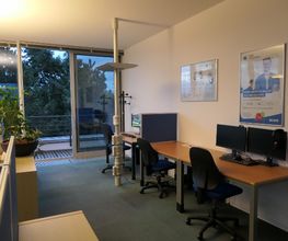 Schulungsraum mit mehreren Arbeitsbereichen, die durch blaue Trennwände voneinander abgegrenzt sind und mit 2 PC-Bildschirmen, Tastatur, Maus und Headset ausgestattet sind. Zugang zu einem Balkon.