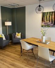 Aufenthaltsraum mit gemütlicher Sitzecke bestehend aus 2 grauen Sesseln und einer modernen Stehlampe. An der Wand hängt ein Bild von einem Wand. Davor steht der große Holztisch mit Stühlen und Blumen.