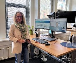 Eine Frau mit Brille und hellbeigem Blazer steht an einem höhenverstellbaren Schreibtisch. Auf dem Tisch stehen ein Monitor, Tastatur, Maus und eine Karaffe. Der Monitor zeigt eine Videokonferenz. Im Hintergrund sind Fenster mit Jalousien.