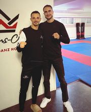 Zwei Athleten stehen in einer Kampfsportschule in sportlicher Kleidung und nehmen eine vorbereitende Kampfhaltung ein. Die Trainingsfläche und Ausrüstung im Hintergrund unterstreichen den professionellen Rahmen.