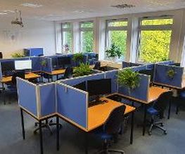 Ein großer Büroraum mit mehreren Arbeitsplätzen, die durch blaue Trennwände abgeteilt sind. Jeder Platz hat einen Bürostuhl, Monitor und Grünpflanzen als Dekoration. Große Fenster lassen viel Tageslicht herein, und draußen sind Bäume sichtbar.