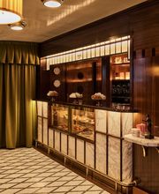 A lit marble pastry display case set into rich wood paneling, featuring warm overhead lighting, green drapes, and a patterned mosaic tile floor.