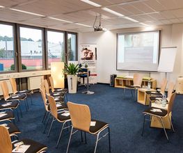 Schulungsraum mit Bestuhlung, an der Wand hängt eine Leinwand, auf die mit einem Beamer eine Präsentation geworfen wird.