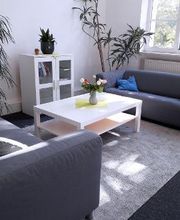 Heller Aufenthaltsraum mit 2 gemütlichen grauen Sofas, einem weißen Beistelltisch auf dem eine Vase mit bunten Blumen steht. sowie ein Schrank und mehrere Zimmerpflanzen.