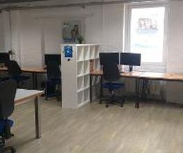 Schulungsraum mit mehreren Arbeitsbereichen, die jeweils mit 2 PC-Bildschirmen, Tastatur, Maus und Headset ausgestattet sind. Weiße Regale dienen als Abtrennung.