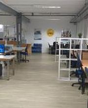 Schulungsraum mit mehreren Arbeitsbereichen, die jeweils mit 2 PC-Bildschirmen, Tastatur, Maus und Headset ausgestattet sind. Weiße Regale dienen als Abtrennung.