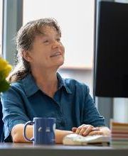 Eine Frau in blauer Bluse sitzt vor einem Monitor und schaut auf den Bildschirm.