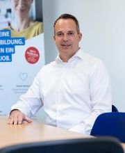 Ein Mann mit kurzen, dunklen Haaren und weißem Hemd sitzt an einem Tisch.