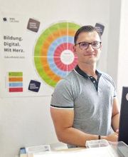 Ein junger Mitarbeiter steht an seinem Schreibtisch und schaut freundlich in die Kamera. Er trägt ein graues T-Shirt und eine Brille. Im Hintergrund hängt ein Poster an der Wand mit der Aufschrift „Bildung. Digital. Mit Herz.“