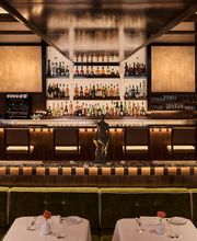 A wide shot of a glowing wood bar lined with spirits bottles and small statues, with green velvet dining booths set with white tablecloths in the foreground.