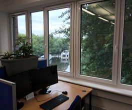Ein Bild von einem Arbeitsbereich neben einem Fenster mit Blick ins Grüne. Der Arbeitsbereich hat blaue Trennwände auf denen ein Blumenkasten angebracht ist sowie 2 Bildschirme, Tastatur und Maus.