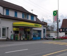 AGROLA Tankstelle in Trübbach. Im Vordergrund Strasse mit Fussgängerstreifen. TAnkstelle mit zwei Tanksäulen an Gebäudefassade. Grosses Totem mit AGROLA Logo und digitaler Preisanzeige.