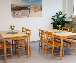 Heller Pausenraum mit hellbraunen Holztischen auf denen gelbe Blumen stehen, Stühlen und einer grünen Zimmerpflanze. An der Wand hängt ein Bild von einem Strand.