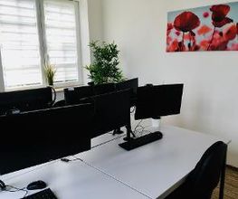 Hell, modern und freundlich gestalteter Prüfungsraum mit 4 weißen Schreibtischen mit Tastaturen und Bildschirmen sowie schwarzen Bürostühlen. Im Hintergrund ein großes Fenster, an der weißen Wand ein längliches Bild mit roten Mohnblumen vor blauem Himmel.