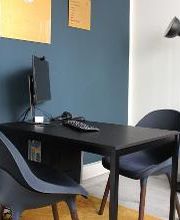 Beratungsraum mit blauer Wand und Plakaten. Davor ein schwarzer Tisch und 2 blaue Sessel. Dahinter eine Stehlampe und ein Regal mit Trockenblumen.