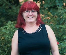 Frau mit rotem Haar und Brille lächelt freundlich. Sie steht vor grünen Pflanzen.
