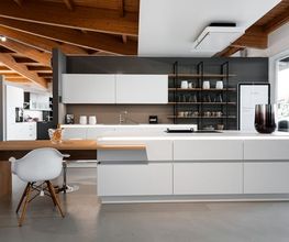 Großzügige, moderne Designküche mit weißen, grifflosen Küchenfronten, einer zentralen Kochinsel und warmen Holzelementen. Die offene Regalwand mit dunklem Metallrahmen, die sichtbare Holzdecke und die hochwertige Ausstattung verbinden minimalistisches Küchendesign mit natürlicher Wohnlichkeit.