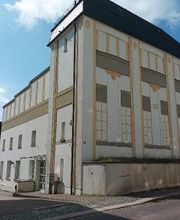 Außenansicht vom Gebäude und Eingang. Das Gebäude hat eine weiße Fassade mit einem gelben Muster.