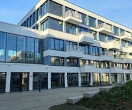 Weißes Gebäude mit mehreren Fenstern und Balkonen. Davor Grünflächen und Pflastersteine