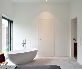 Reduziertes, modernes Interieur mit klarer Formsprache: Ein freistehendes, skulpturales Bad im Schlafzimmer verleiht dem Raum eine besondere architektonische Note. Die bodentiefen Fenster bringen Tageslicht in die minimalistisch gestaltete Umgebung, während warme Lichtakzente eine wohnliche Atmosphäre schaffen. Die Kombination aus Funktion und Design macht den Raum zu einem Statement für zeitgenössisches Wohnen.