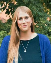 Frau mit blondem Haar steht vor grünem Baum und lächelt freundlich.