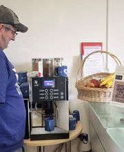 Ein Mann steht vor einem Kaffeevollautomat und macht sich einen Kaffee. Daneben ein Obstkorb sowie ein Aufsteller mit einem QR Code und ein kleiner Ventilator im Hintergrund.