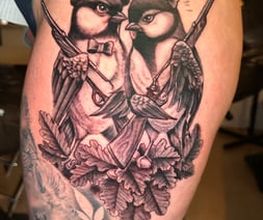 Detailreiches Black-and-Grey-Tattoo mit zwei verliebten Vögeln, Gewehren, Herzen und Eichenlaub auf dem Oberschenkel.