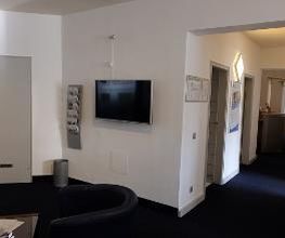 Empfangsbereich und Flur mit Sesseln und einem TV. Daneben hängt ein Flyerhalter.