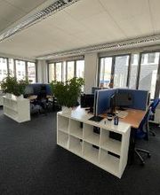 Schulungsraum mit mehreren Arbeitsbereichen, die mit blauen Trennwänden voneinander getrennt sind. Auf jedem Tisch befinden sich 2 Bildschirme, eine Tastatur, Maus und ein Headset. Es befinden sich 2 weiße Regale und mehrere Zimmerpflanzen in dem Raum.