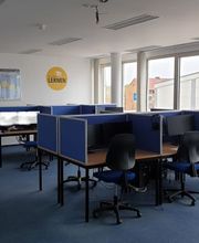Schulungsraum mit mehreren Arbeitsbereichen, die durch blaue Trennwände voneinander abgegrenzt sind und mit 2 PC-Bildschirmen, Tastatur, Maus und Headset ausgestattet sind sowie blauen Bürostühlen.