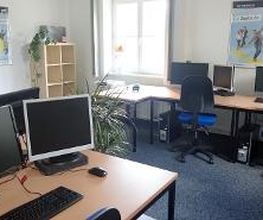 Schulungsraum mit 4 Arbeitsplätzen jeweils mit 2 Bildschirmen, einem Bürostuhl, Tastatur und Maus. An den Wänden hängen Plakate. In der Ecke steht ein Regal mit einer Pflanze.