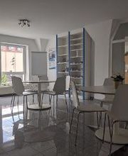 Aufenthaltsraum mit 2 Tischen und jeweils 5 Stühlen. Im Hintergrund sind 2 große Fenster und ein Bücherregal. Links steht eine Zimmerpflanze.