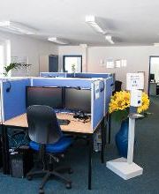 Dieses Bild zeigt einen Schulungsraum. Zu sehen sind Schreibtische, welche jeweils mit zwei Bildschirmen, einer Tastatur und Maus sowie einem Headset ausgestattet sind. In dem Raum befindet sich eine große Vase mit Sonnenblumen.