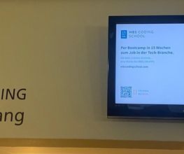 Teilnehmerinforamtionen auf einem Fernseher, der an einer Wand im Eingangsbereich hängt. Daneben steht geschrieben WBS TRAINING Empfang mit Richtungspfeilen.