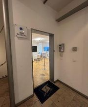 Geöffnete Eingangstür im Inneren des Gebäudes. Auf dem Boden liegt eine blaue Fußmatte mit dem WBS Logo aufgedruckt und links daneben hängt ein Schild auf dem das WBS TRAINING Logo und die Aufschrift WBS TRAINING zu lesen ist.