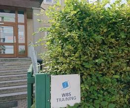Logo von WBS TRAINING am Eingang in der Hecke, im Hintergrund die Treppe und die Eingangstür.