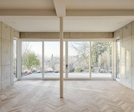 Offener Wohnraum mit raumhoher Verglasung, Sichtbetonwänden und Fischgrätparkett. Architektur, Licht und Landschaft treten in einen ruhigen, atmosphärischen Dialog.