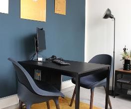 Beratungsraum mit blauer Wand und Plakaten. Davor ein schwarzer Tisch und 2 blaue Sessel. Dahinter eine Stehlampe und ein Regal mit Trockenblumen.