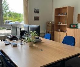 Ein Büro mit 2 Arbeitsplätzen bestehend aus jeweils 2 PC-Bildschirmen, Tastatur, Maus und kleinen Topfpflanzen. Im Hintergrund ein Schrank mit Regal sowie eine Pinnwand.