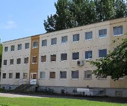 Außenansicht des Schulgebäudes an einem sonnigen Tag. Das Gebäude hat eine helle Fassade und ist von Bäumen umgeben.