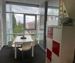 Ein heller Pausenraum mit einem weißen Tisch und vier weißen Stühlen. Rechts steht ein Regal mit roten und weißen Fächern, darauf ein Modell eines Segelboots. Große Fenster bieten einen Blick auf Bäume und umliegende Gebäude.