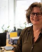 Eine Frau mit lockigem Haar, Brille und dunkelgrüner Bluse lächelt in die Kamera. Im Hintergrund sind ein Schreibtisch mit Monitor, Pflanzen und verschwommen Fenster mit Blick auf ein Gebäude zu sehen. Der Raum ist hell und freundlich eingerichtet.