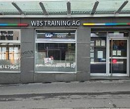 Haupteingang mit zwei großen Fenstern neben der Tür. Auf der grauen Fassade ist der Schriftzug WBS TRAINING AG angebracht.