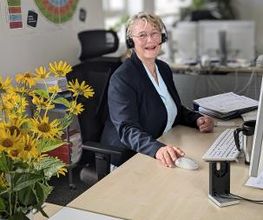 Frau sitzt am Arbeitsplatz mit Tastatur und Maus. Neben ihr ein gelber Blumenstrauß, hinter ihr ein großes Plakat