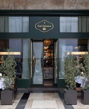 Exterior of Sant Ambroeus with a deep green façade, gold-trimmed logo above the entrance, sheer-curtained windows, and outdoor tables framed by potted olive trees.
